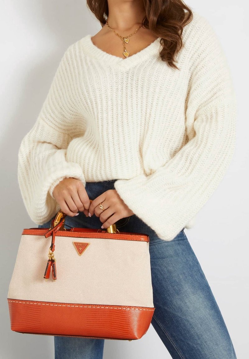 Femme en pull en maille blanche et jeans bleus tenant un sac à main Guess beige et orange avec des poignées dorées et un détail pompon.