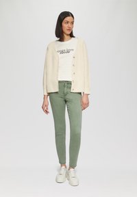 Kremowy kardigan z dzianiny z guzikami, w paski biało-beżowa koszula, dopasowane oliwkowe spodnie i białe sportowe trampki na platformie. Minimalistyczny styl.