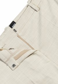 Beige räätälöidyt housut, jotka on valmistettu tekstuurista kankaasta. Housuissa on vetoketjukiinnitys, vyölenkit ja logotagi. Korostuksena on erivärinen sisätasku.
