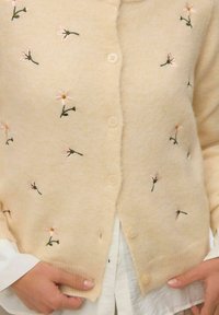 Béžový kardigan s malou růžovou květinovou výšivkou, nošený přes bílou košili, ruce drží okraje kardiganu u knoflíků.