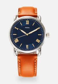 Montre Fossil avec un boîtier rond en acier argenté, un cadran bleu marine avec des chiffres romains dorés et un bracelet en cuir marron avec des détails de couture.