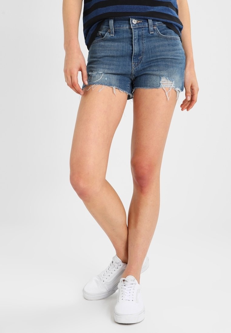 Pantalones cortos de mezclilla, azul claro, desgastados con dobladillos deshilachados. Combinados con una camisa a rayas azul marino y zapatillas blancas. Ajuste y estilo casual.