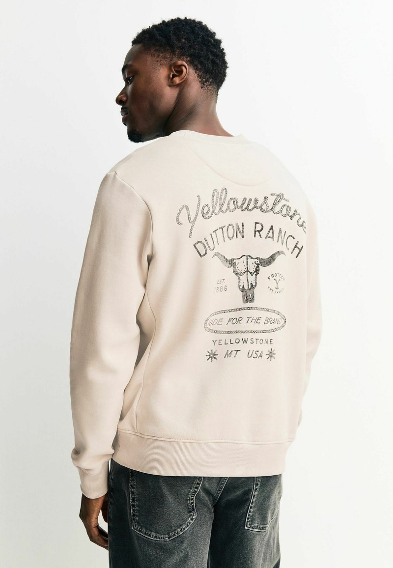 Sweat-shirt beige avec un col rond et des manches longues. Présente un motif graphique de crâne et du texte au dos en couleurs contrastantes.