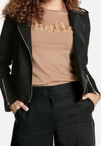 Veste en simili cuir noir avec quincaillerie argentée sur une chemise beige ornée d'une broderie texturée "Hello", accompagnée d'un pantalon noir.