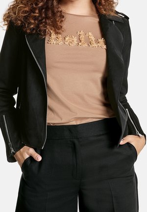 Veste en simili cuir noir avec quincaillerie argentée sur une chemise beige ornée d'une broderie texturée "Hello", accompagnée d'un pantalon noir.