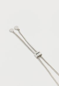 Ασημένιο ρυθμιζόμενο βραχιόλι αλυσίδας με συρόμενο κούμπωμα χαραγμένο με "DKNY" και δύο επίπεδα στρογγυλά καπάκια στις άκρες της αλυσίδας σε λευκό φόντο.