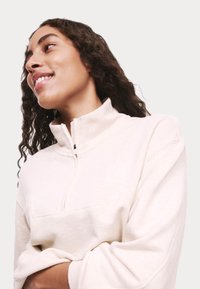 Crèmekleurige, ritsloze pullover met een hoge kraag en gebreide stof. Beschikt over een subtiele naden aan de voorkant en ontspannen mouwen.