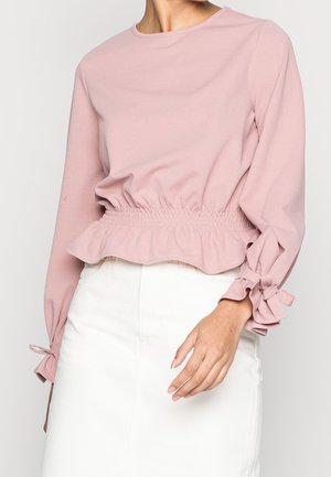 Femme portant un haut à manches longues rose clair avec taille élastique et détails de nœuds aux poignets, associé à une jupe blanche.