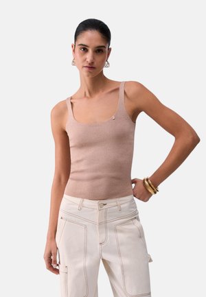 Žena v bezovom tielku bez rukávov a svetlých vysokých nohaviciach, s zlatými kruhovými náušnicami a náramkami, stojaca pred jednoduchým pozadím.