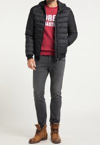 Schwarze Steppjacke mit Kapuze, Reißverschluss und elastischen Bündchen, getragen über einem roten Sweatshirt und grauen Jeans, kombiniert mit braunen Stiefeln.