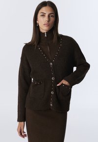 Bruine gebreide cardigan met decoratieve knopen, twee voorkruiden, ribgebreide manchetten en een V-hals ontwerp boven een bijpassende ribgebreide jurk.