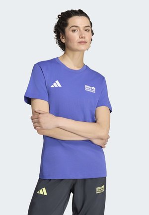 Persoon met kort krullend haar die een blauw BMW Berlin Marathon t-shirt en zwarte short met Adidas-logo's draagt, staand met gekruiste armen.