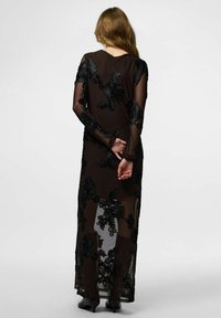 Robe maxi noire transparente ornée de broderies florales, manches longues et silhouette ajustée. Le dos est légèrement ouvert avec un décolleté arrondi.