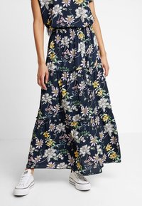 Femme portant une robe maxi florale bleu marine avec des baskets blanches, se tenant devant un fond blanc uni.