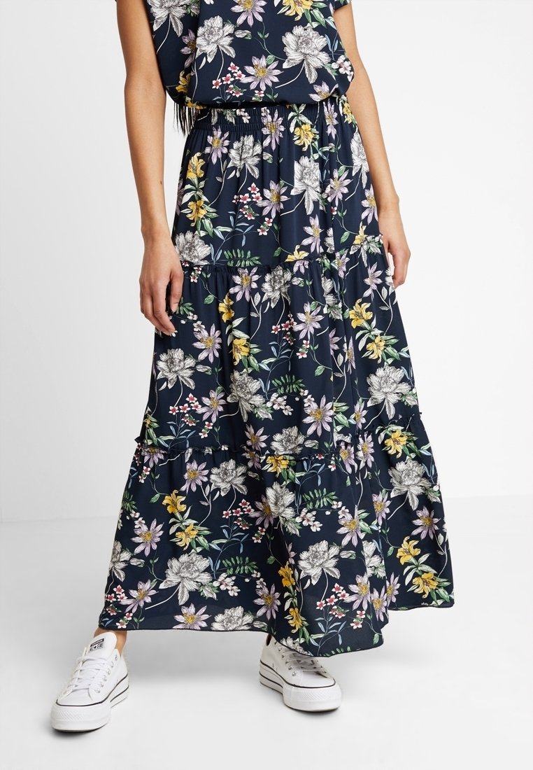 Femme portant une robe maxi florale bleu marine avec des baskets blanches, se tenant devant un fond blanc uni.