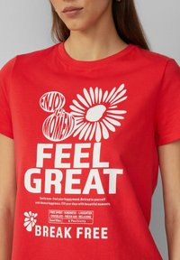 Rode katoenen T-shirt met witte tekst en bloemenillustraties. De tekst bevat "GENIET VAN HET MOMENT," "VOEL JE GOED," en "BREK LOS." Zachte textuur.