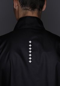 Veste noire avec une texture lisse, un col, et une ligne verticale de petites croix blanches en décoration au dos.