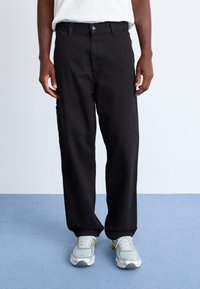 Black denim pants with a relaxed fit, featuring a button fly, front and back pockets, and a smooth texture. Worn with gray sneakers.