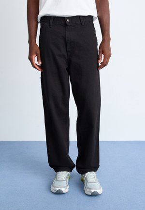 Black denim pants with a relaxed fit, featuring a button fly, front and back pockets, and a smooth texture. Worn with gray sneakers.