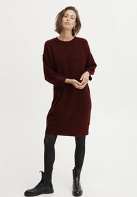 Abito maglione bordeaux realizzato in morbido tessuto a maglia, con una vestibilità comoda, maniche lunghe e orlo al ginocchio. Indossato con stivaletti neri alla caviglia.
