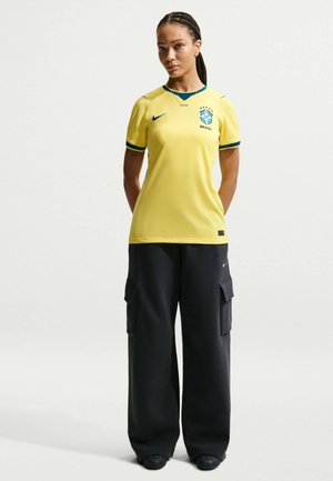 Vrouw staand met handen achter de rug, gekleed in een geel Braziliaans voetbalshirt en zwarte broek met wijde pijpen tegen een effen achtergrond.