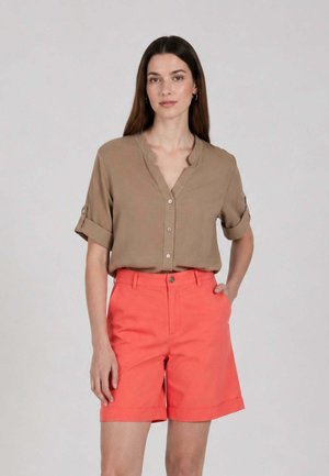 Donna con capelli lunghi e castani che indossa una camicia beige a maniche corte abbottonata e pantaloncini corallo al ginocchio, in piedi davanti a uno sfondo semplice.