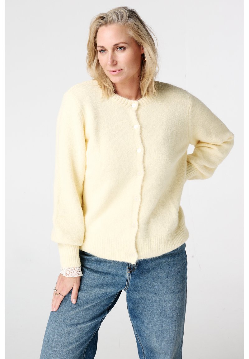 Lichtgele, zachte cardigan met een ronde hals, lange mouwen en knopen aan de voorkant. Gecombineerd met blauwe denim, voorzien van een subtiel kantdetail bij de manchetten.