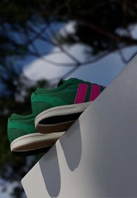 Zelené sieťované tenisky s ružovými pruhmi, bielymi podrážkami a hnedými akcentmi, umiestnené na rímske proti rozmazanému vonkajšiemu pozadiu.