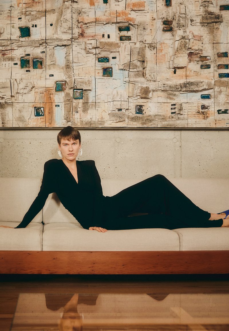 Zwart aansluitend langslavende jurk, persoon liggend op een lichtbeige bank met een houten basis. Achtergrond met abstracte wandkunst in gedempte tinten.