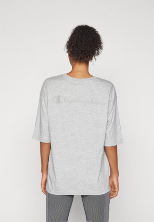 Personne aux cheveux bouclés portant un t-shirt Champion ample gris clair, de dos, debout devant un fond clair uni.