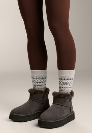Szürke velúr bokacsizma bolyhos szegéllyel, fehér mintás zoknival párosítva. A lábak barna leggingsben vannak, semleges háttér előtt.