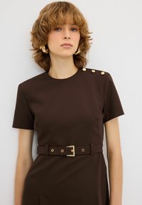 Bruine aansluitende jurk met korte mouwen, ronde halslijn, gouden knoopaccenten op de schouder en een bijpassende riem met een gouden gesp.