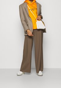 Blazer beige sobre una sudadera naranja, combinado con pantalones anchos marrones y zapatillas blancas. Se lleva un pequeño bolso blanco cruzado sobre el cuerpo.