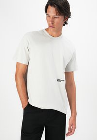 Ung mand iført en lys grå Karl Lagerfeld T-shirt og sorte bukser, poserer med den ene hånd i lommen og ser til venstre.