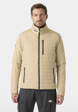 Hombre con chaqueta acolchada beige con cremalleras delanteras y pantalones cargo negros con logo en el bolsillo izquierdo, de pie frente a un fondo liso.