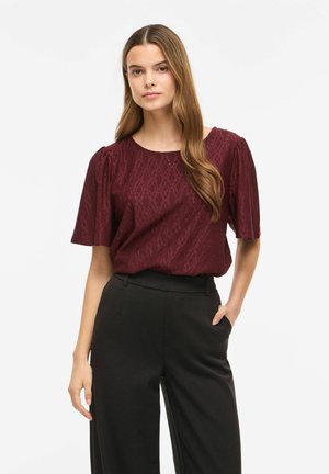 Jeune femme aux longs cheveux bruns portant un chemisier bordeaux à motifs à manches courtes et un pantalon noir taille haute, debout avec une main dans la poche.