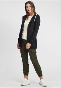 Felpa nera con zip e cordini regolabili, abbinata a pantaloni cargo verde oliva. Il modello indossa sneakers bianche con dettagli gialli.
