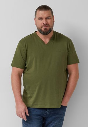 Mężczyzna z brodą i związanymi włosami, ubrany w oliwkowozieloną koszulkę z dekoltem w serek i niebieskie dżinsy, stojący na tle jednolitego szarego tła.