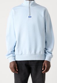 Sudadera con cremallera de color azul claro, con un alto cuello, puños y dobladillo acanalados. Cuenta con un pequeño logo azul en el pecho. Textura de tela suave.