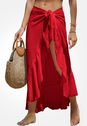 Roter Wickelrock mit einer Bindeschließe an der Taille, ausgestattet mit einem rüschengesäumten Saum und einem hohen Schlitz. Kombiniert mit einer handgewebten Strohtasche.