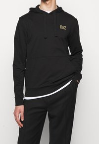 Svart hoodie med en framstående känguruficka, prydd med en liten guldfärgad logga. Den har en dragsko vid huven och ribbade muddarna för en åtsittande passform.