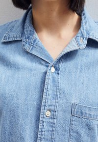 Ljusblå denimskjorta med krage, fyra knappar och en framficka. Tyget är strukturerat med en lätt sliten utseende.