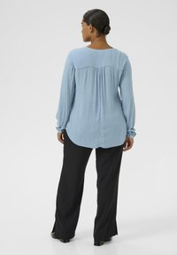 Blouse bleu clair à manches longues avec un col rond et un détail froncé à l'arrière, assortie à un pantalon noir et des chaussures à talons.