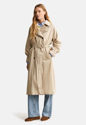 Γυναίκα που φοράει μπεζ καμπαρντίνα με ζώνη πάνω από μπλε πουκάμισο και φαρδιά τζιν, σετ με καφέ σουέντ παπούτσια, στέκεται με το ένα χέρι στην τσέπη.