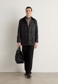 Veste imperméable noire avec plusieurs poches, une fermeture à boutons et un sac noir texturé. Portée par-dessus une chemise à boutons marron clair et un pantalon noir.