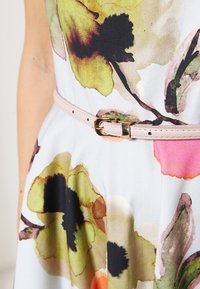 Femme portant une robe blanche ornée de grands motifs floraux jaunes, noirs et roses, cintrée à la taille par une fine ceinture beige avec une boucle dorée.