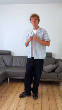 Camisa de mangas curtas com riscas brancas por cima de uma t-shirt branca simples, combinada com calças largas pretas e sapatos pretos, num ambiente interno minimalista.