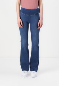 Jeans in denim blu a zampa d'elefante con vita alta e dettagli di cucitura sottili; indossati con un top rosa chiaro e sneakers bianche.