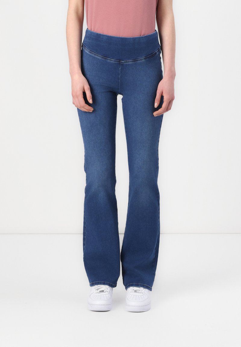 Jeans in denim blu a zampa d'elefante con vita alta e dettagli di cucitura sottili; indossati con un top rosa chiaro e sneakers bianche.