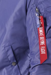 Ljubičasta najlonska bomber jakna s prednjim dugmetom, zippered džepom i istaknutom crvenom oznakom od tkanine s natpisom "UKLONITE PRIJE LETA."